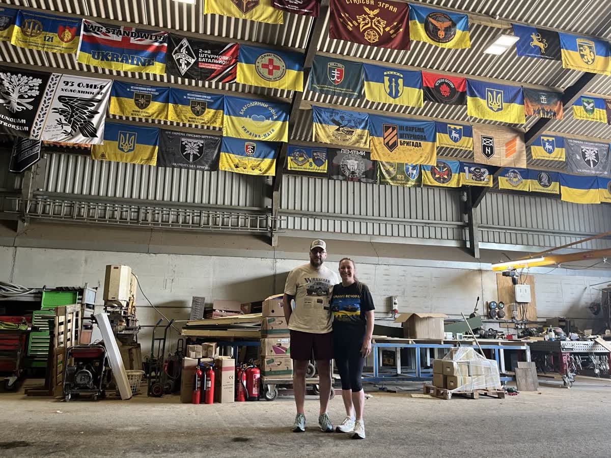 Two crew members standing in the loading barn beneath dozens of Ukrainian brigade banners hung along the roofline