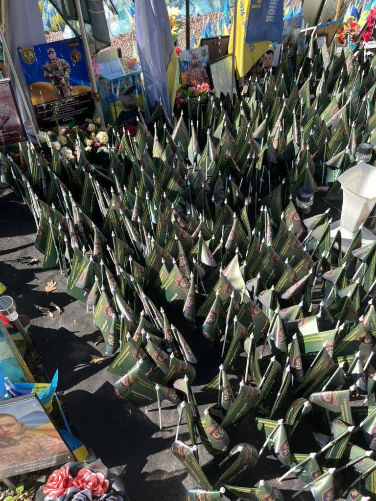 A dense field of small memorial flags bearing unit insignia, set among portraits and flowers on Maidan in Kyiv