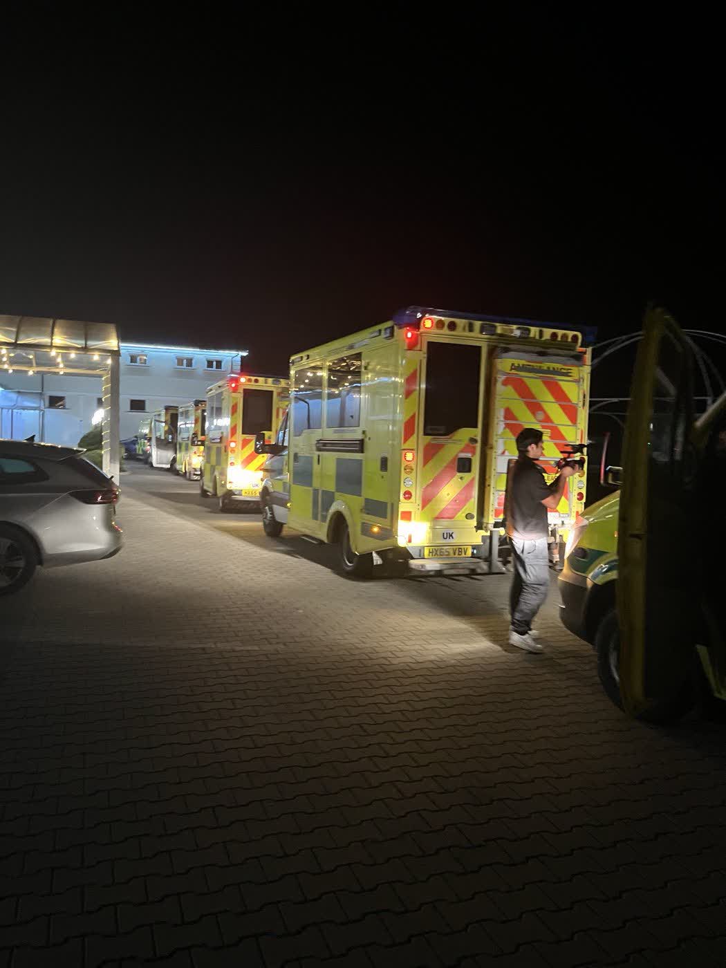 Convoy ambulances parked in a car park at night with tail lights on, a crew member walking between them