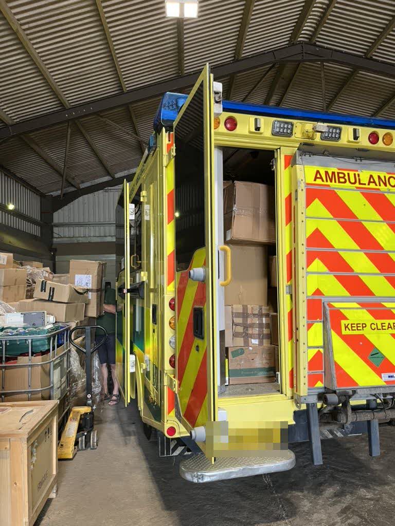 An ambulance with rear doors open, packed to the roof with boxes of aid, surrounded by supplies in the warehouse